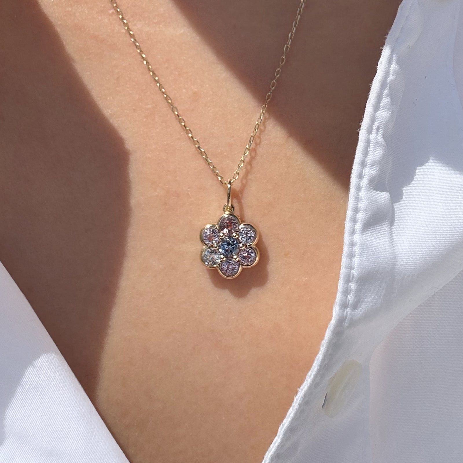 A gold flower-shaped pendant necklace with clear and light blue gemstones, worn on a model's neck over a white collared shirt.