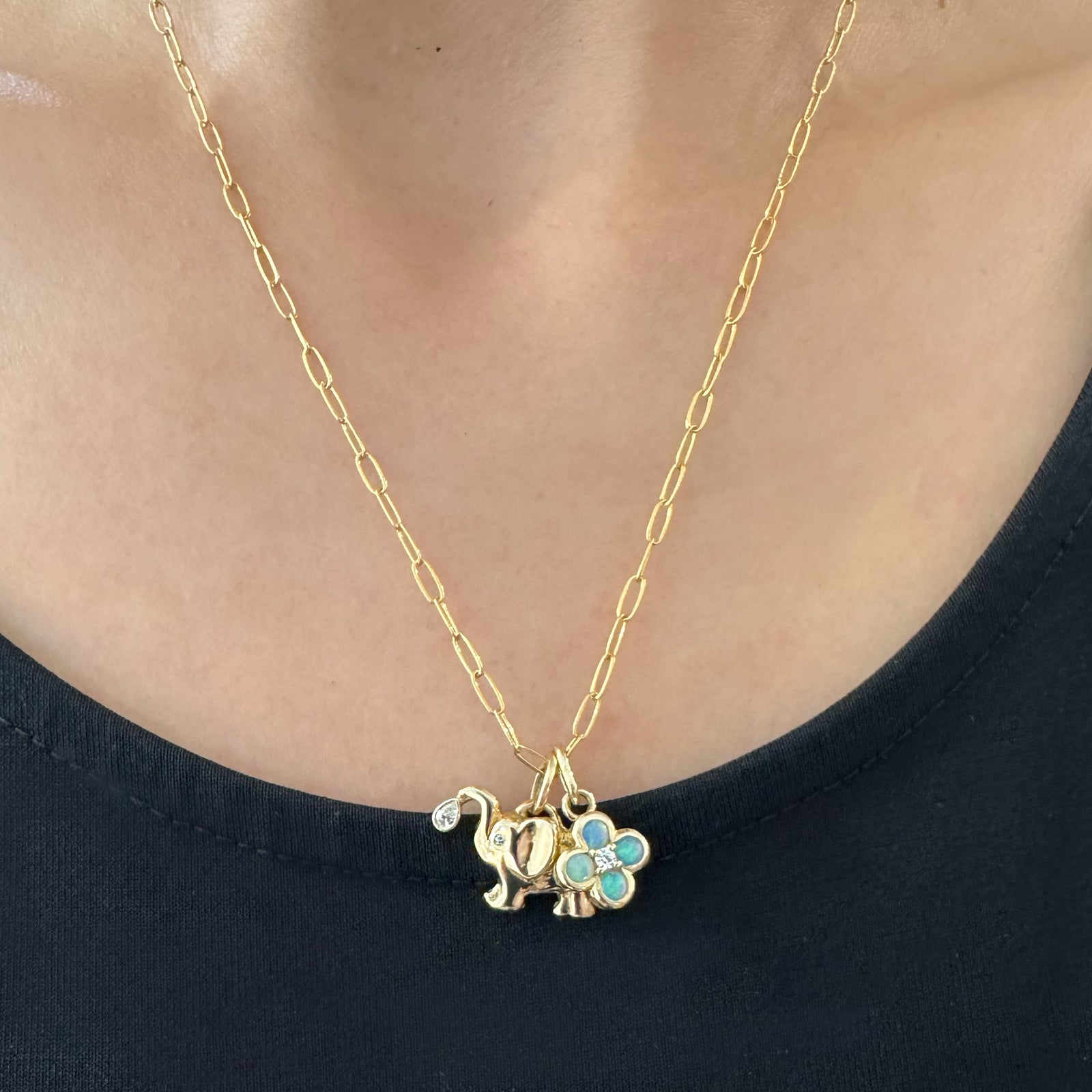 A close-up of a person wearing a gold chain necklace with a small elephant charm and a four-leaf clover charm embellished with opal stones.