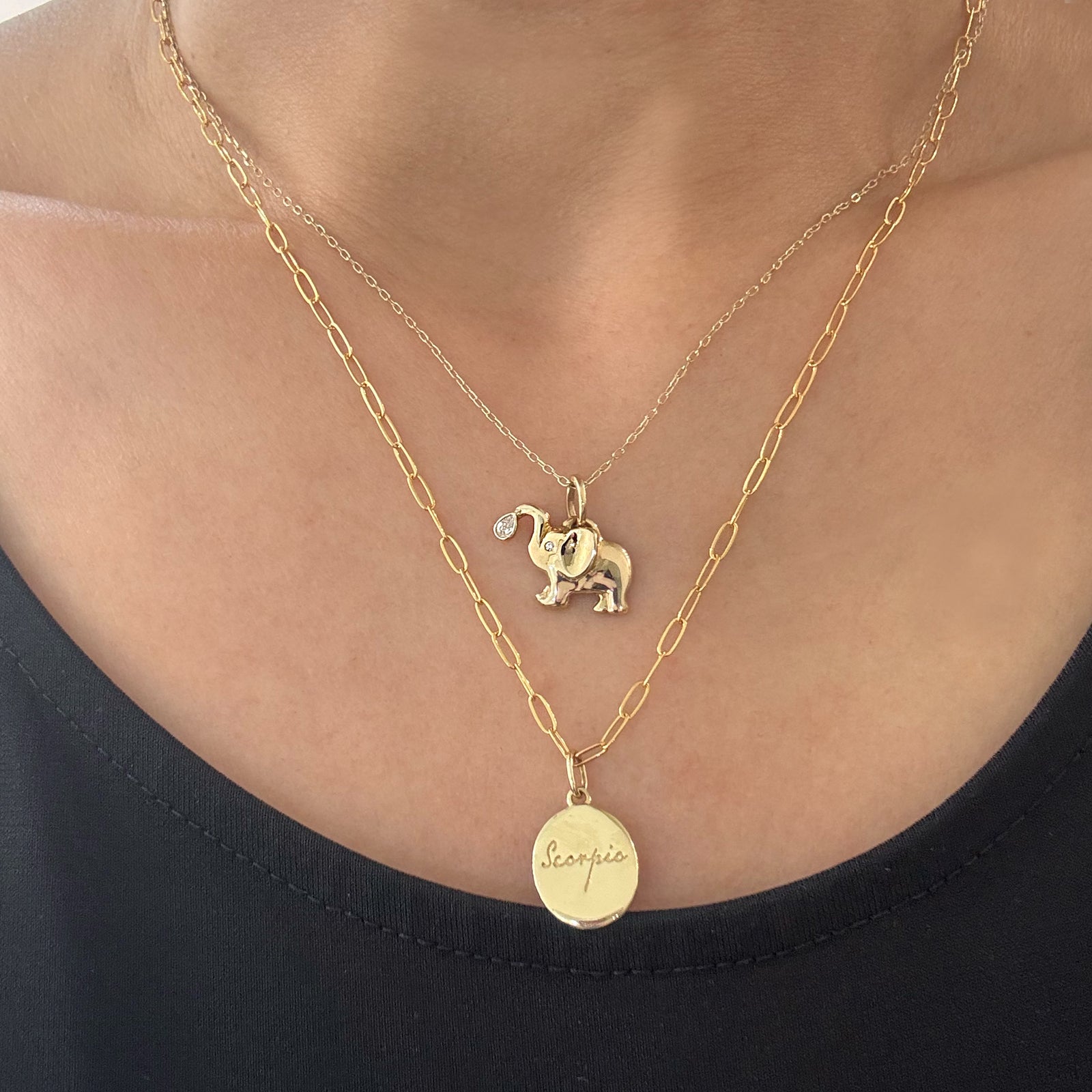 A close-up of a person wearing a layered gold necklace set. The top layer is a delicate chain with a small elephant charm, and the bottom layer is a slightly thicker chain with a oval pendant engraved with 'scorpio'.