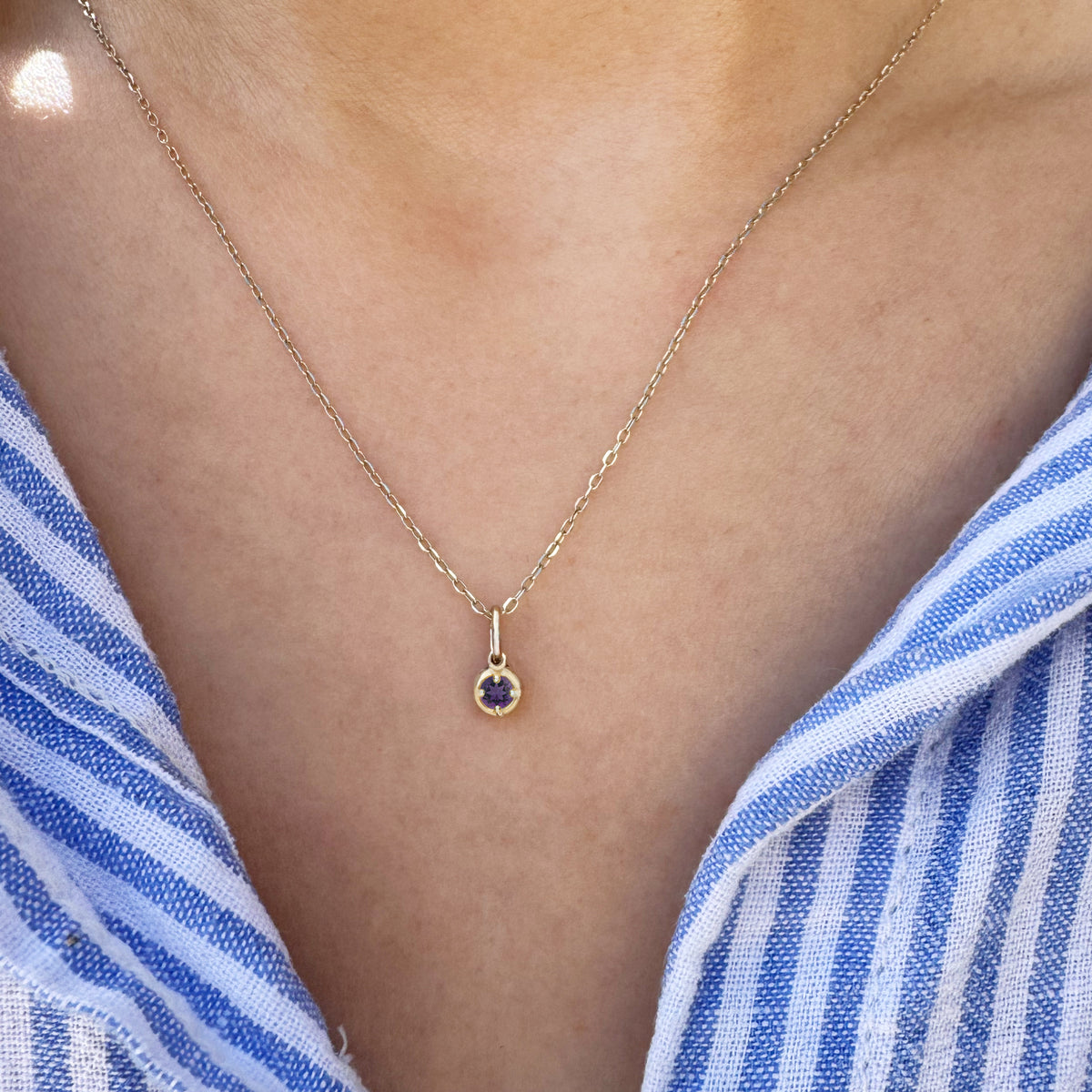 A delicate gold chain necklace with a small round purple gemstone pendant, worn on a model's neck over a blue and white striped shirt.