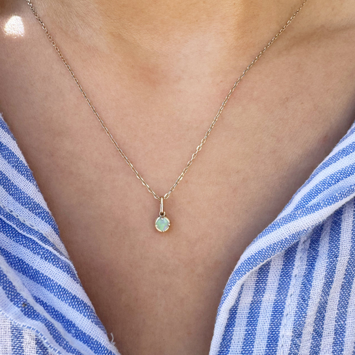A delicate gold chain necklace with a small round-set  opal  pendant, worn on a model's neck over a blue and white striped shirt.