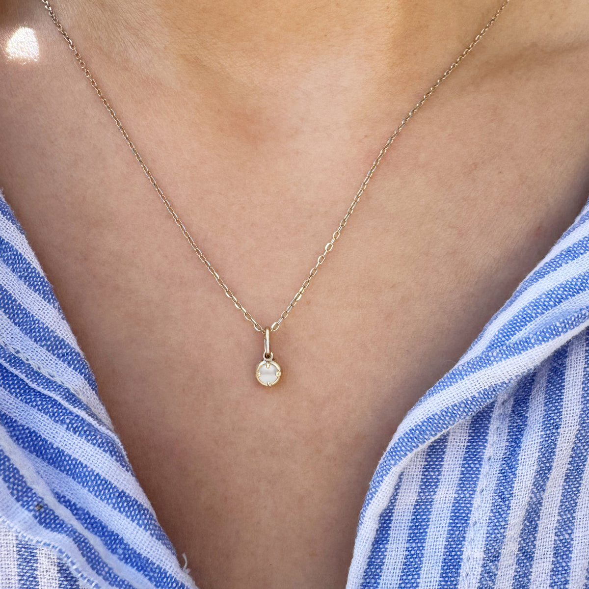 A delicate gold chain necklace with a small round pearl pendant, worn on a model's neck over a blue and white striped shirt.