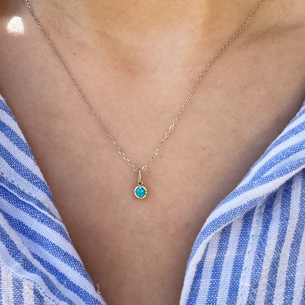 A delicate gold chain necklace with a small bezel-set turquoise pendant, worn on a model's neck over a blue and white striped shirt.