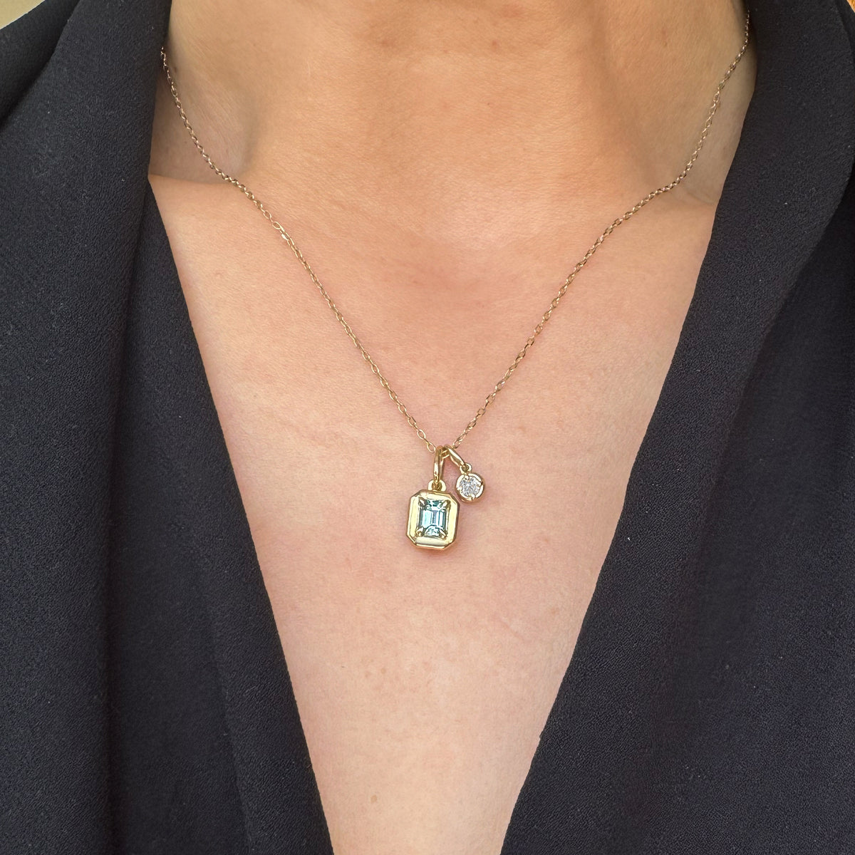 A close-up of a person wearing a delicate gold chain necklace with a small rectangular light blue gemstone pendant and a tiny clear stone accent, styled with a black blazer.