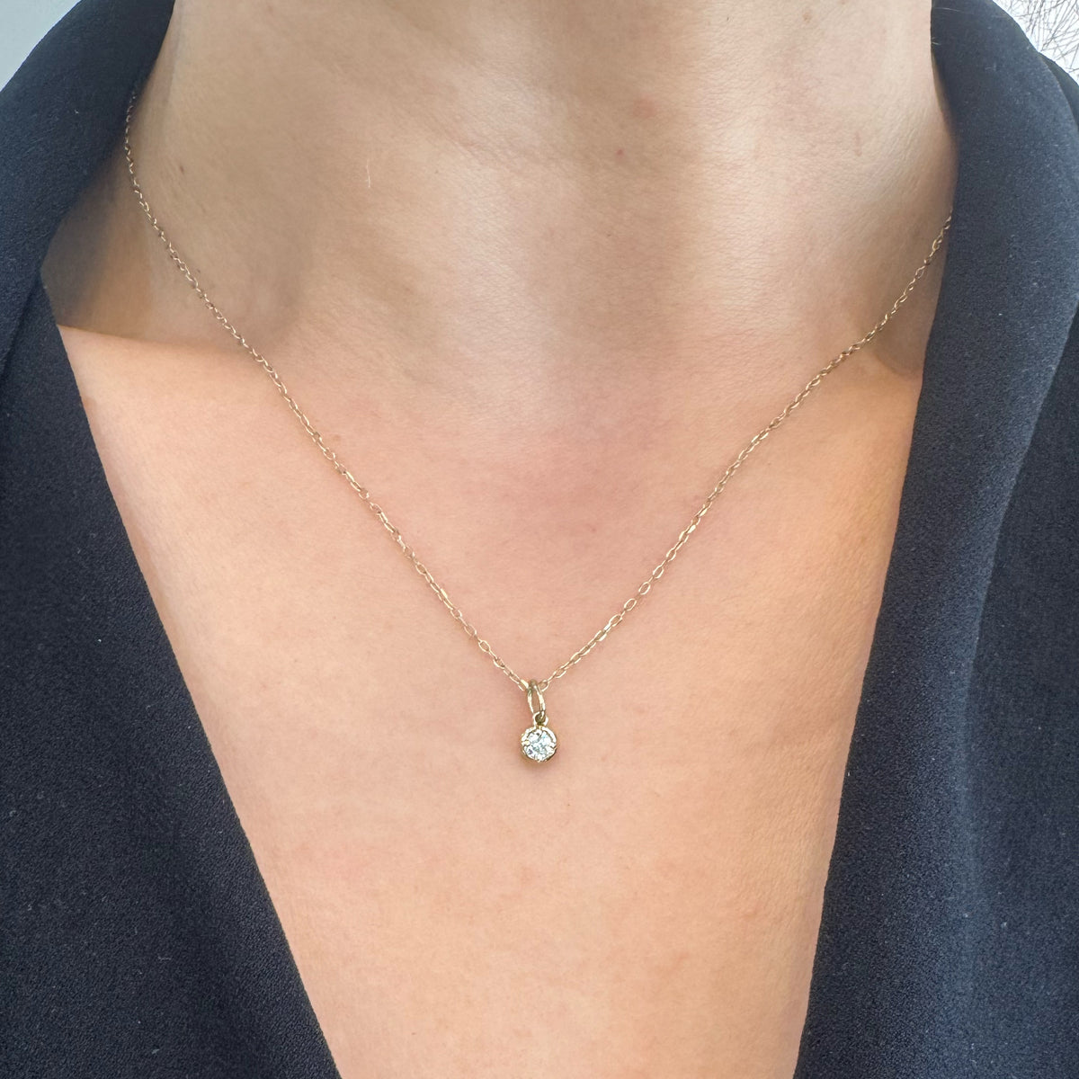 A delicate gold chain necklace with a small diamond gemstone pendant, shown worn on a model's neck over a black V-neck top.