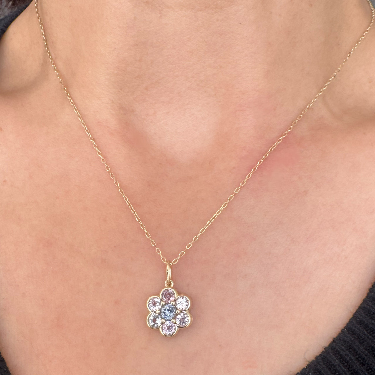 A close-up of a person's neck and chest wearing a delicate gold chain necklace with a flower-shaped pendant. The pendant features clear and light pink gemstones in a gold setting.