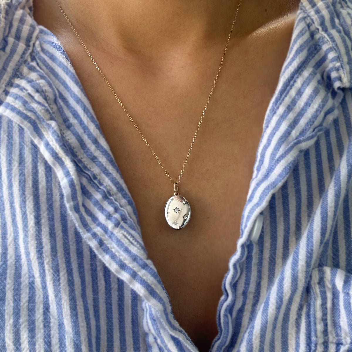 A close-up of a person wearing a delicate gold chain necklace with an oval silver pendant engraved with the star patterens styled with a blue and white striped shirt.
