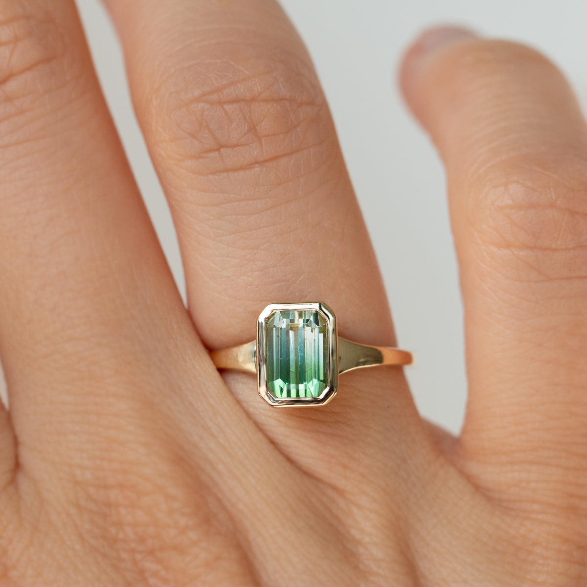 A close-up of a hand wearing a gold ring with a rectangular emerald-cut green gemstone.