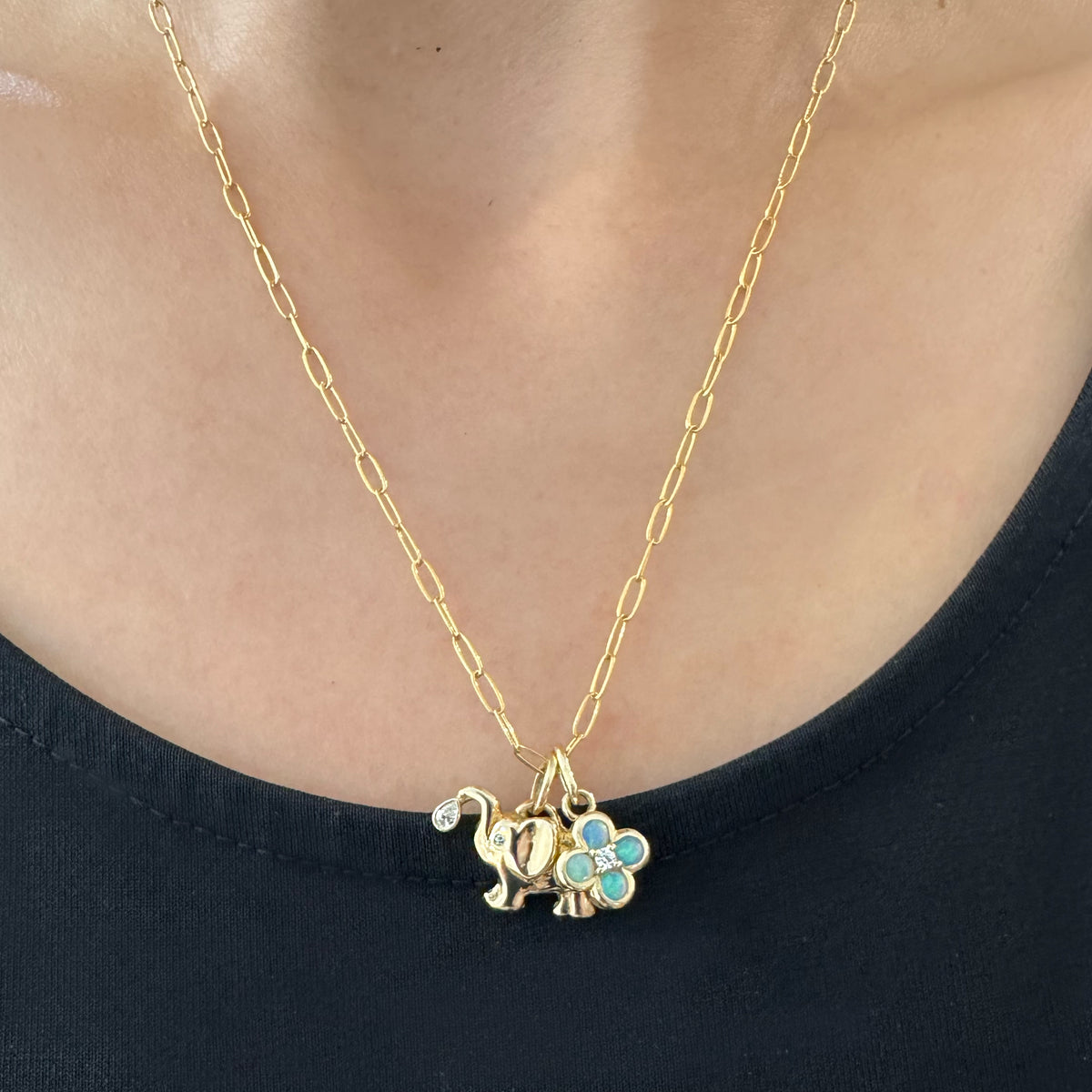 A close-up of a person wearing a gold chain necklace with a small elephant charm and a four-leaf clover charm embellished with opal stones.