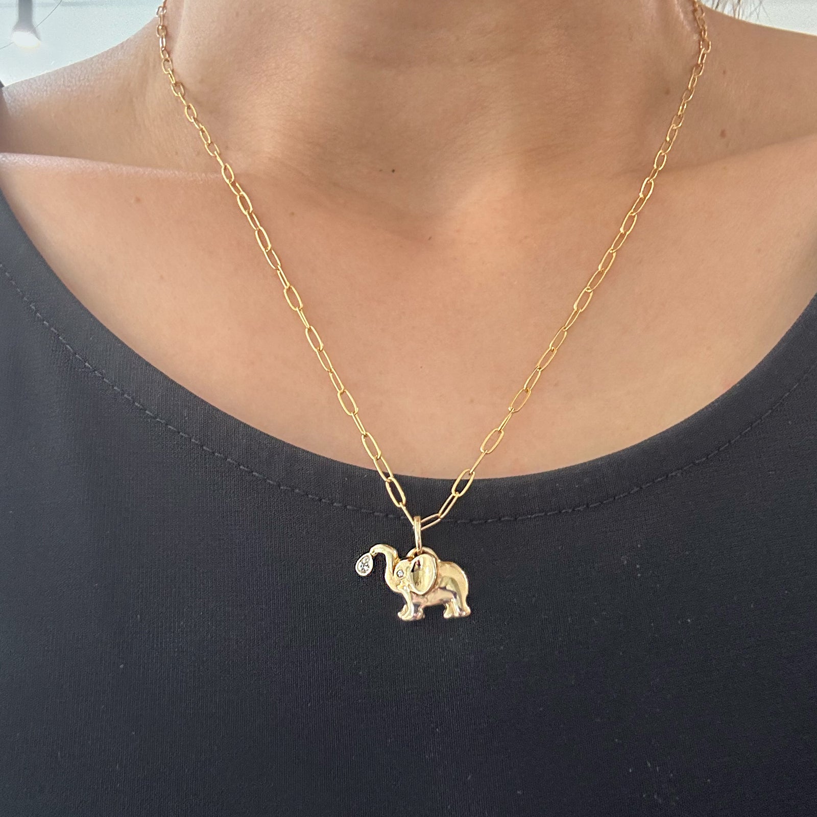 A gold paperclip chain necklace with a small elephant pendant featuring a diamond eye, worn by a model wearing a black top.