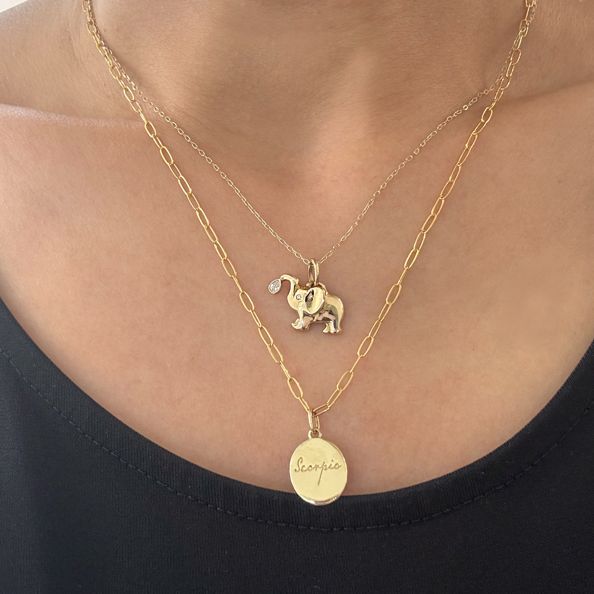 A close-up of a person wearing a layered gold necklace set. The top layer is a delicate chain with a small elephant charm, and the bottom layer is a slightly thicker chain with a oval pendant engraved with 'scorpio'.