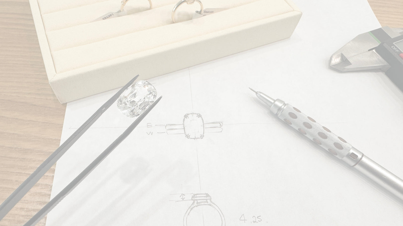 A close-up of a ring design and setting process on a wooden table, featuring a cream-colored jewelry box with two finished rings, a hand holding tweezers with a clear gemstone, a silver mechanical pencil, and a caliper over a sketch of a ring on white paper.