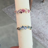 A Gold ring with pink gemstone on top and a blue gemstones ring on the bottom of a white rod against a gray background.  