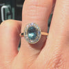 teal sapphire and white diamond halo ring on a hand against a outside background. 
