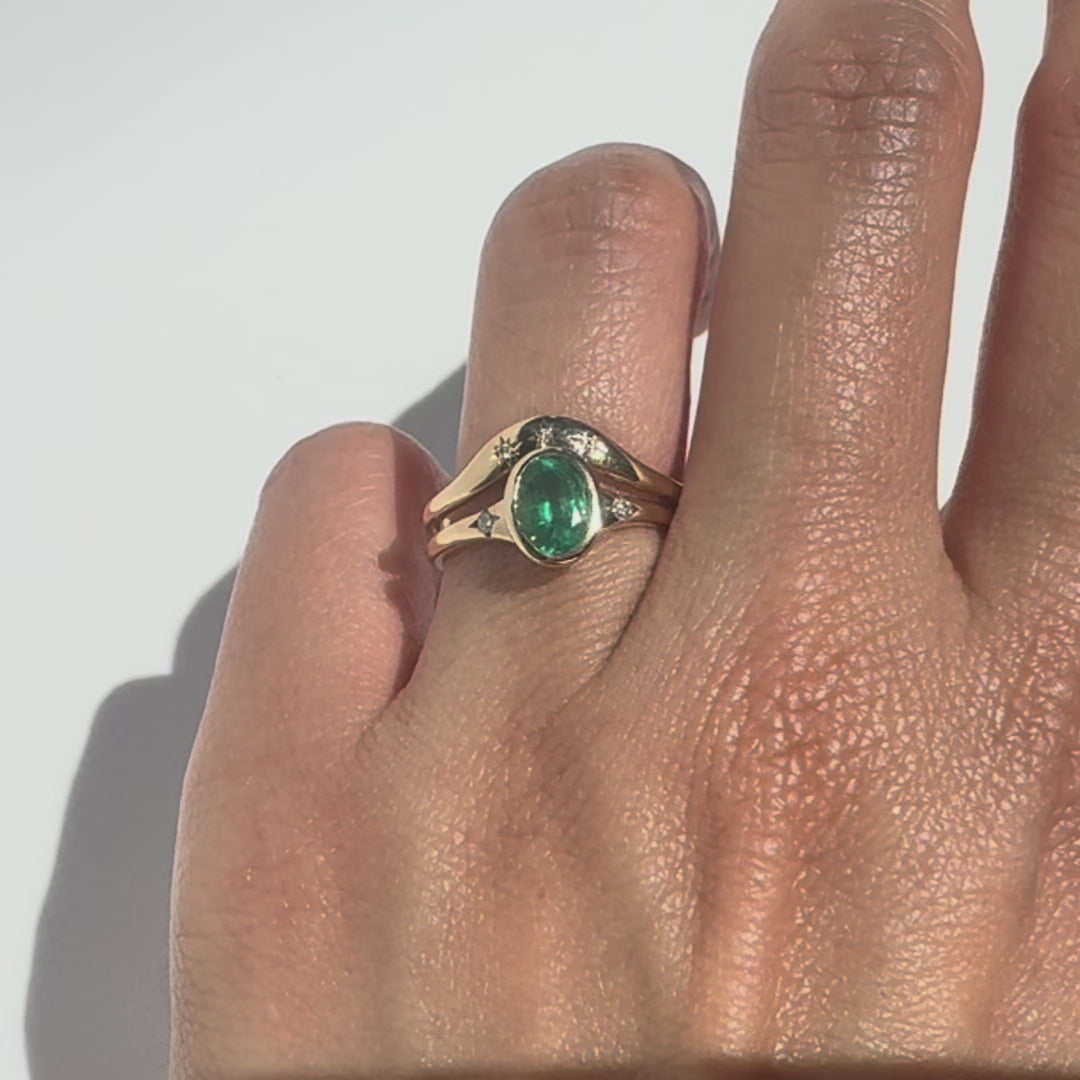hand wearing an oval emerald set gold ring and a contour gold band accented with thre diamonds on white background. 
