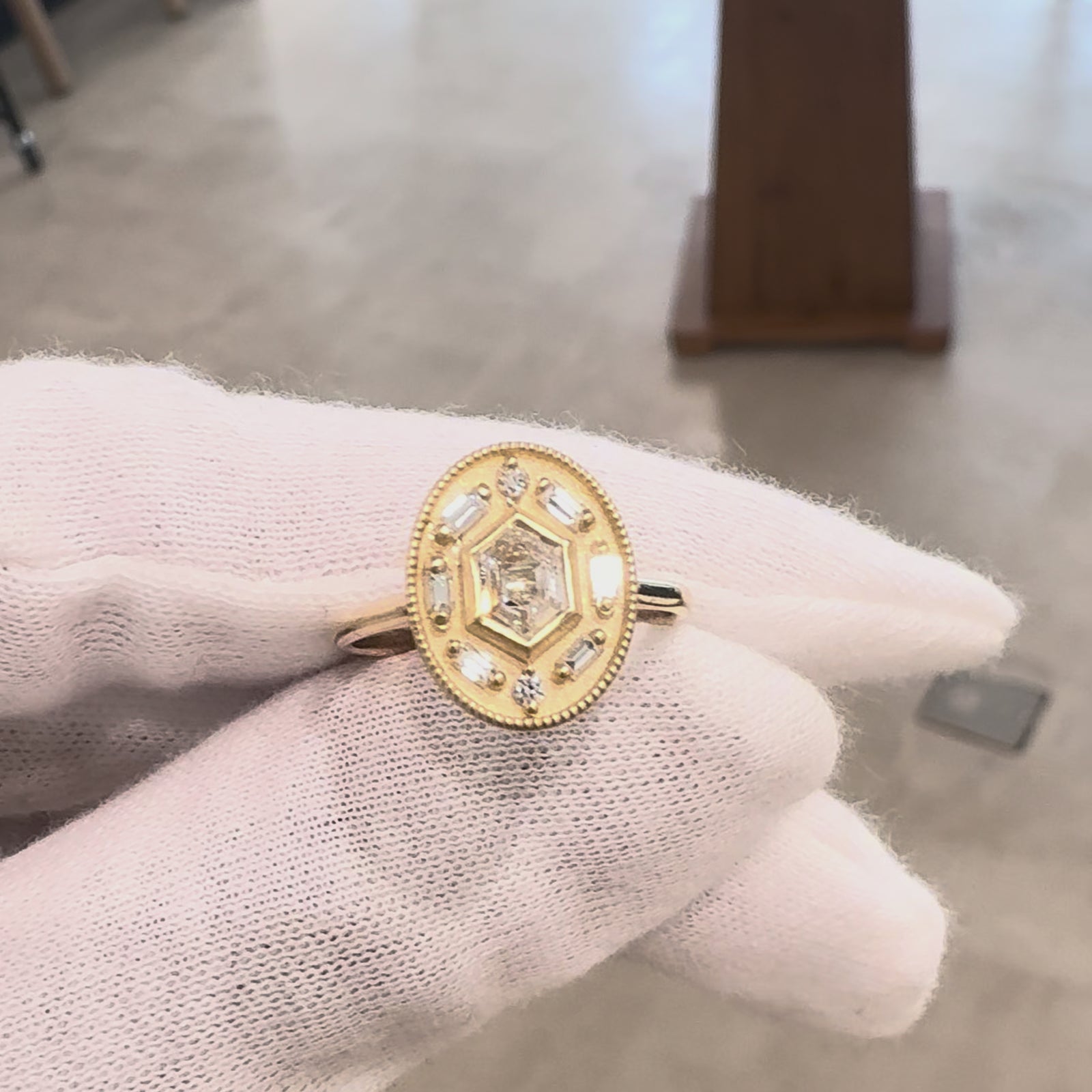 Oval ring with a hexagon shaped diamond in the center held in a hand in a white glove against a gray background. 