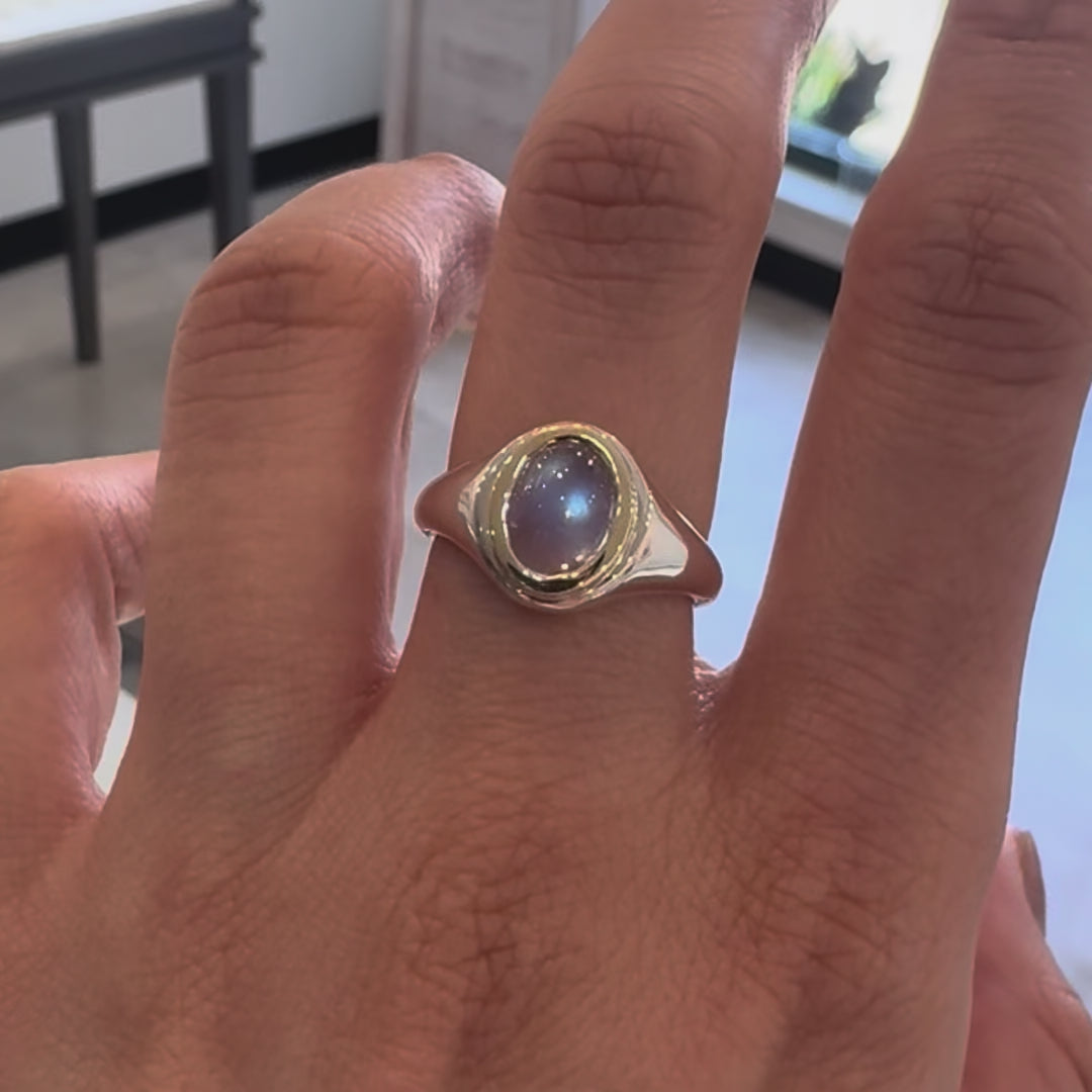 Ring with a blue moonstone and gold bezel setting on a silver band on a hand in an indoor background. 