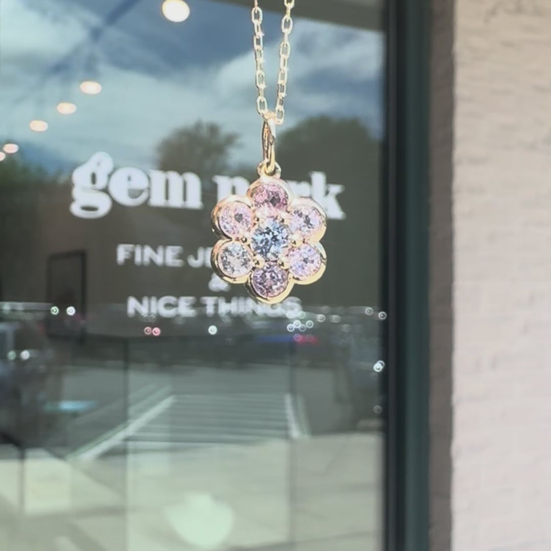 A gold flower-shaped pendant necklace with clear, pink and blue sapphire against a glass window