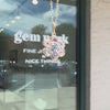 A gold flower-shaped pendant necklace with clear, pink and blue sapphire against a glass window