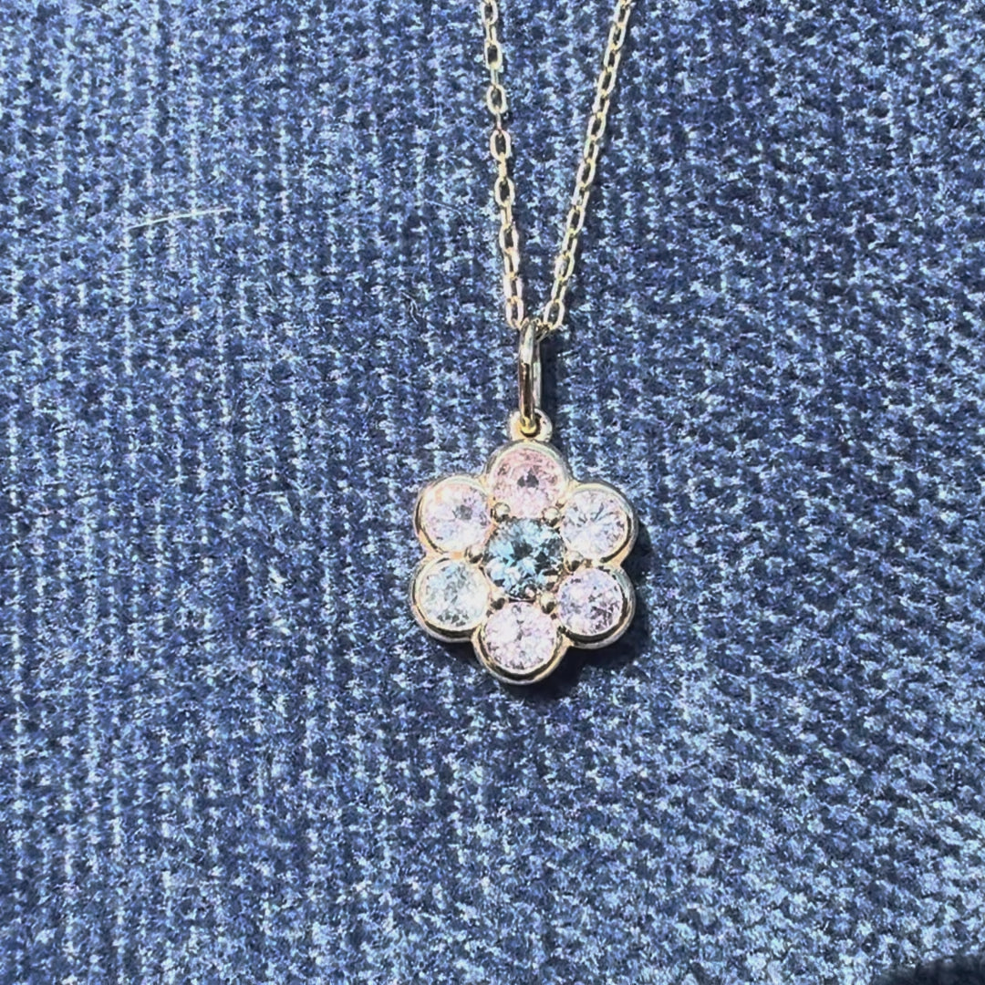 A gold flower-shaped charm pendant featuring a central round blue gemstone surrounded by six smaller round gemstones in pale pink, clear, and light blue, set in a polished gold bezel on a blue background.