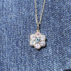 A gold flower-shaped charm pendant featuring a central round blue gemstone surrounded by six smaller round gemstones in pale pink, clear, and light blue, set in a polished gold bezel on a blue background.